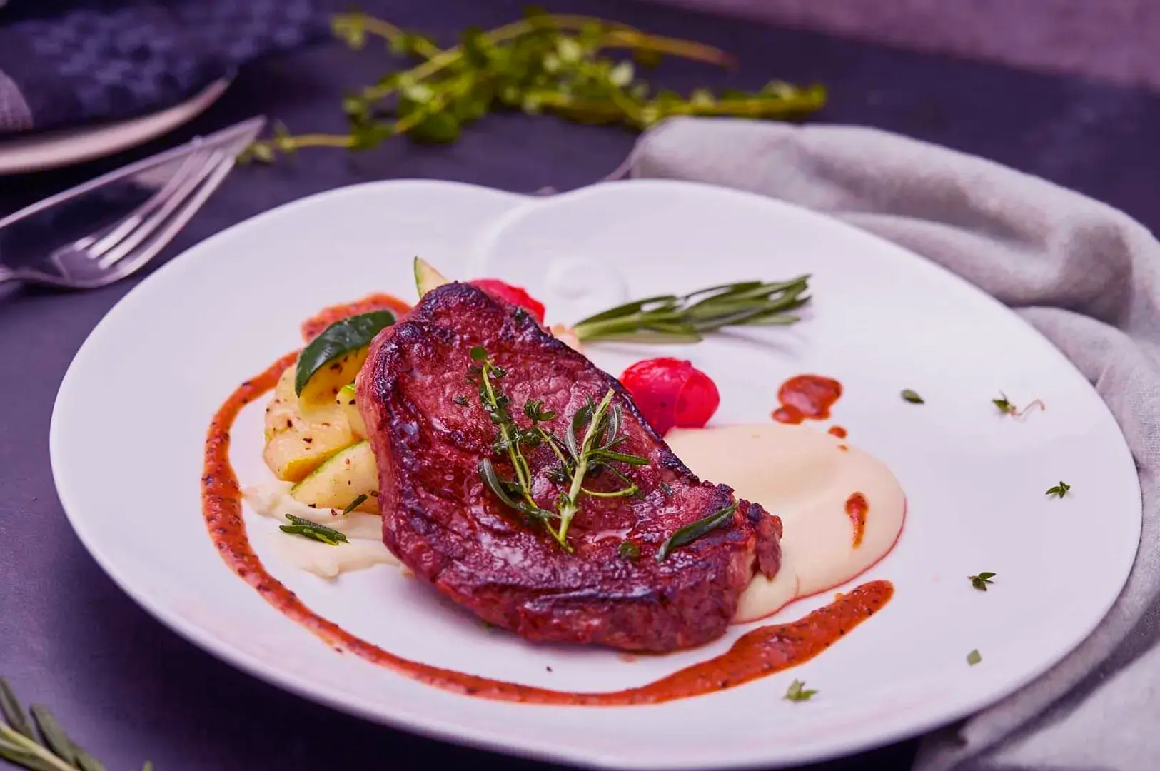 Grilled steak garnished with herbs on a white plate, accompanied by mashed potatoes, zucchini, cherry tomatoes, and a drizzle of sauce. Elegant composition.