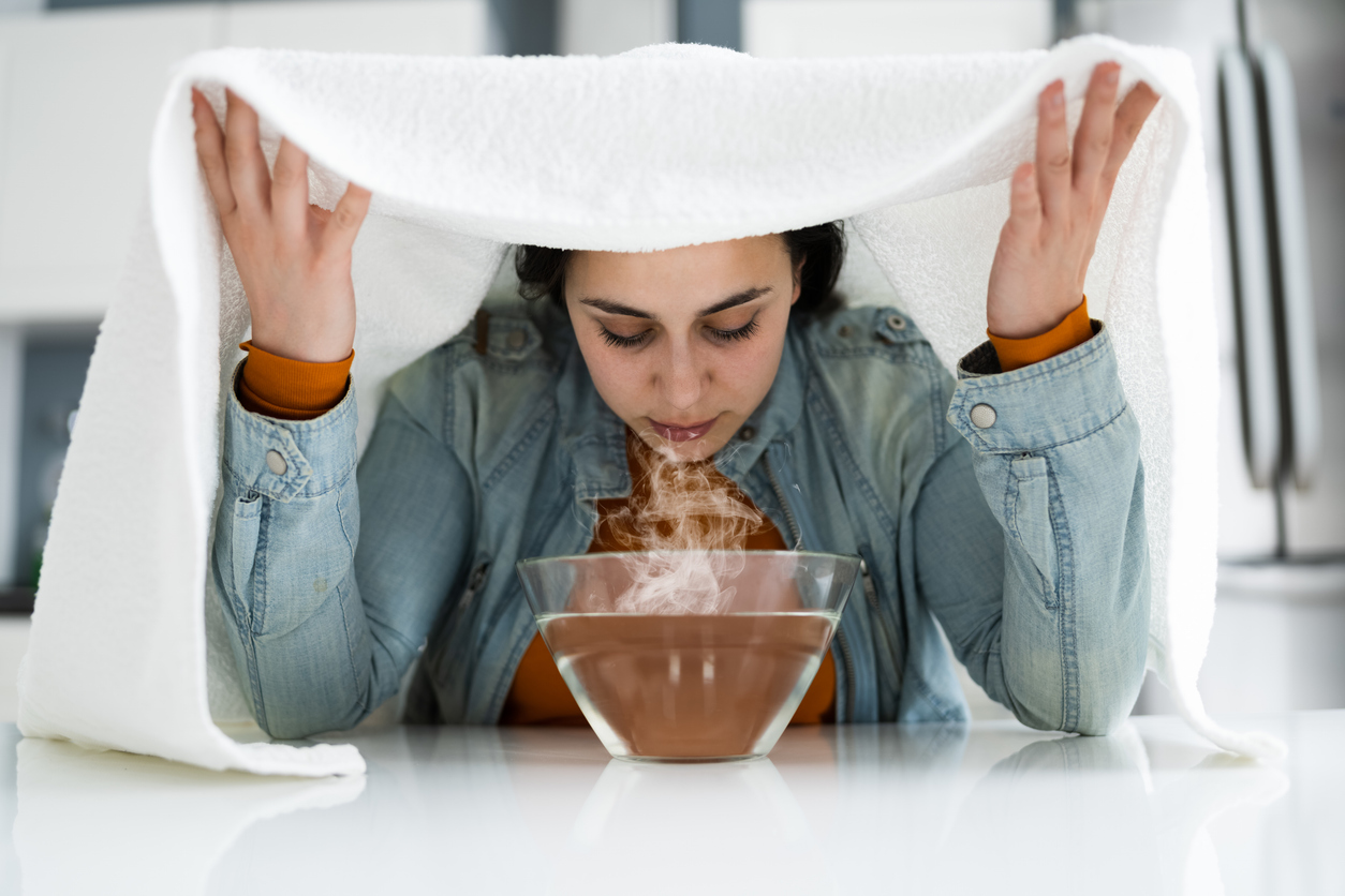 An image of a woman with a towel draped over her head breathing steam out of a bowl.