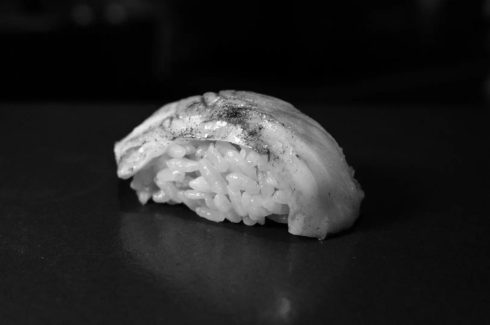 Close-up of a nigiri sushi piece topped with a thin slice of fish on a dark surface.