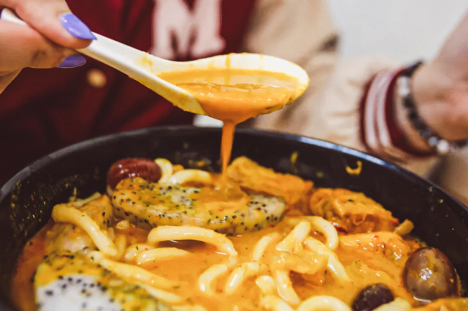 Fruit Juice Curry noodle soup with thick noodles, rice cakes, sausages, tofu, and vegetables in creamy orange broth, spoon lifting rich sauce.