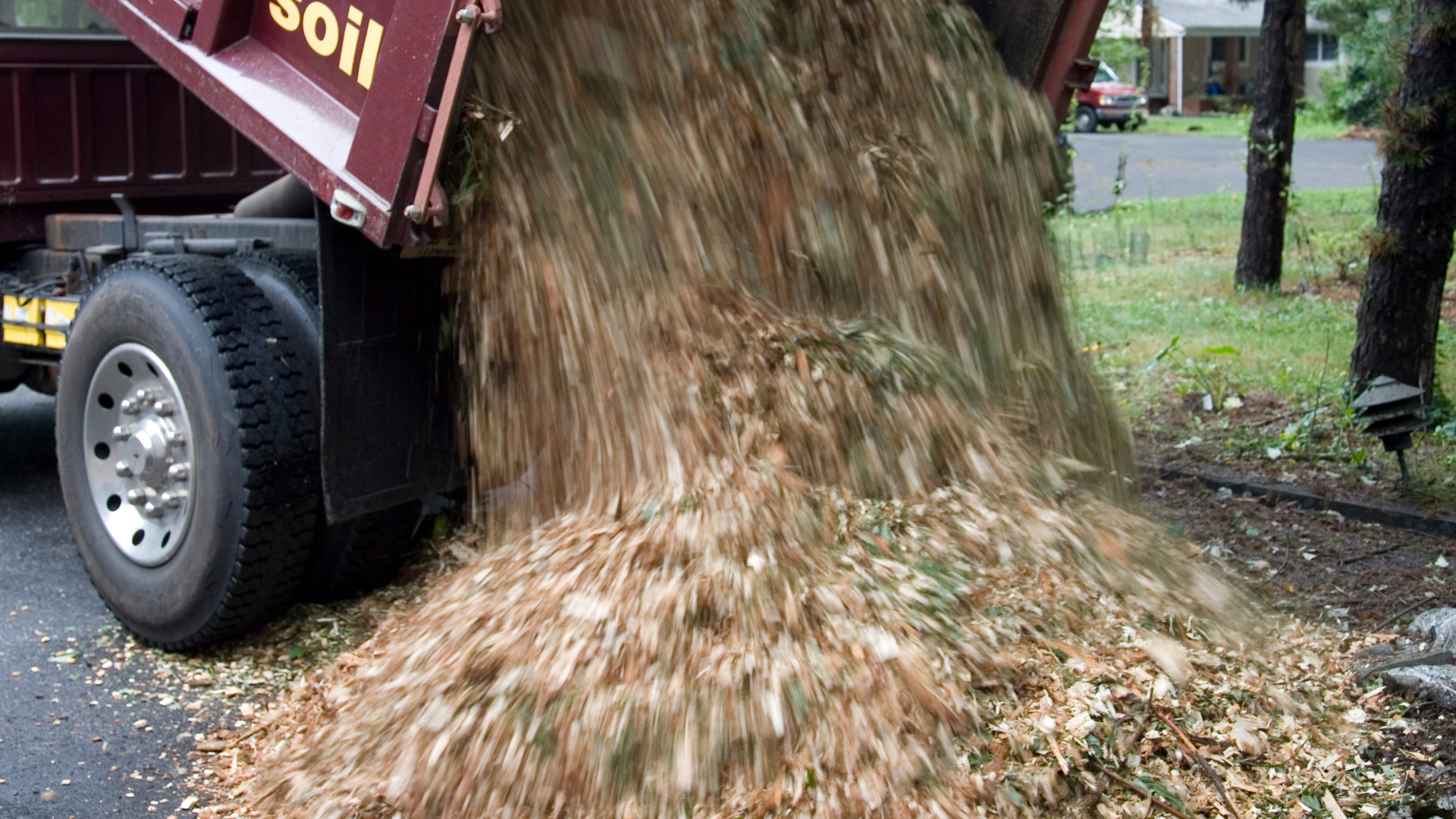 A truck dumping off a load of wood chips to one side of a property.