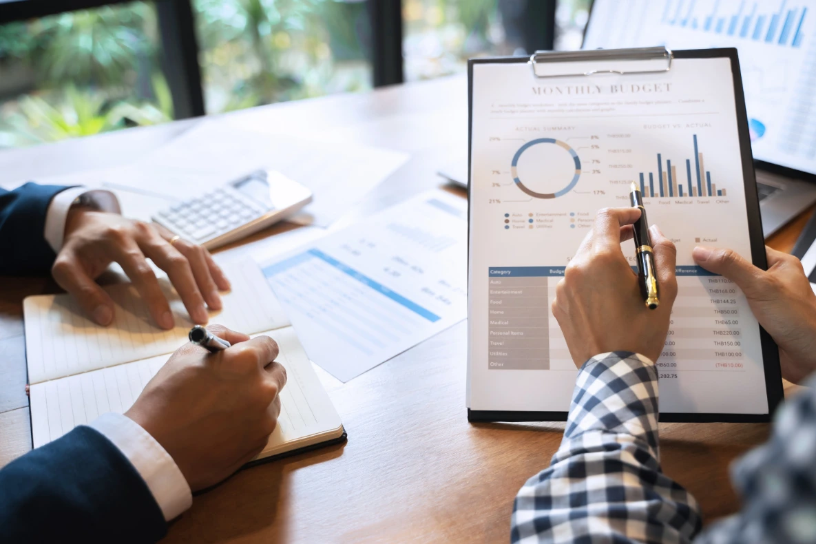 Two individuals are reviewing financial documents, with one person writing in a notebook and the other pointing to a chart on a clipboard that displays a monthly budget.