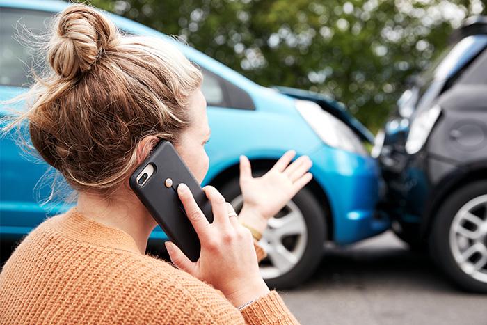 A woman seen from behind, wearing an orange sweater with her hair up, talking on a cell phone at a car accident scene with two collided vehicles visible in the background, depicting the immediate steps to take after a Texas car crash.