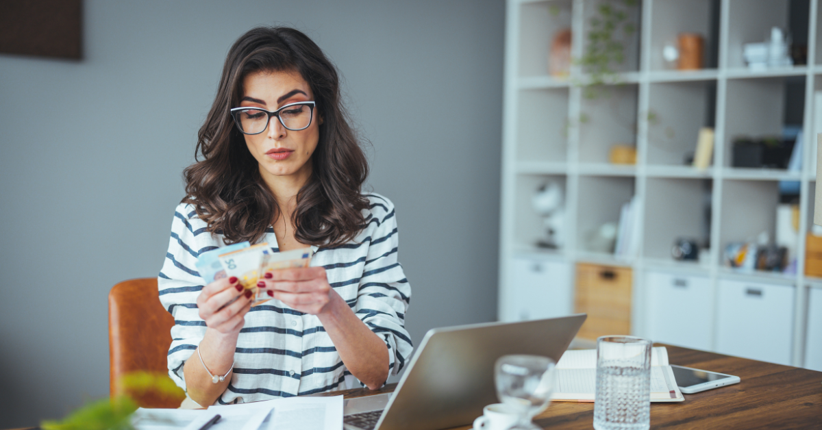 A focused woman counting cash at a desk with a laptop; wondering how much tax you pay on OnlyFans income?