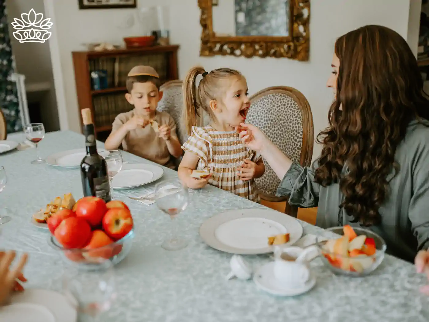 A mother feeding her daughter at a festive Rosh Hashanah table, with another child eating in the background - Fabulous Flowers and Gifts, Rosh Hashanah Flowers Collection.