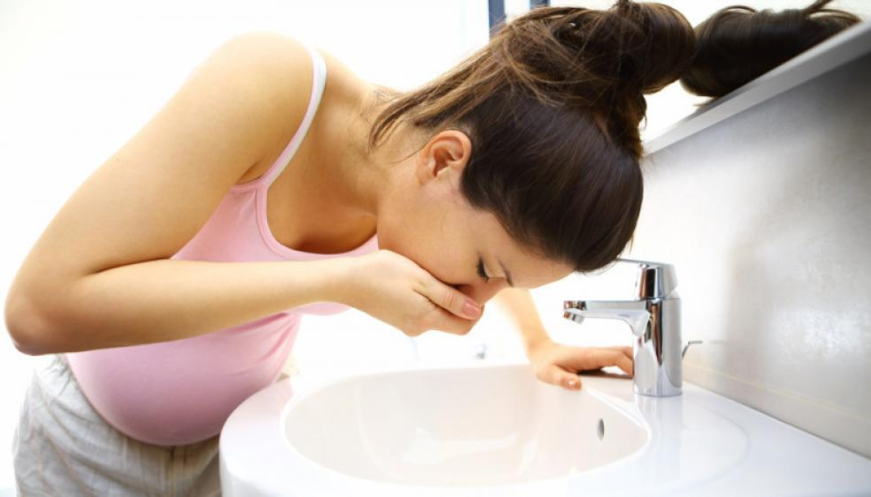 Kvalme gravid – Derfor føler du deg uvel (og hva som faktisk hjelper) 9 Gravid kvinne med håret i hestehale lener seg over vasken og dekker munnen, trolig på grunn av kvalme.