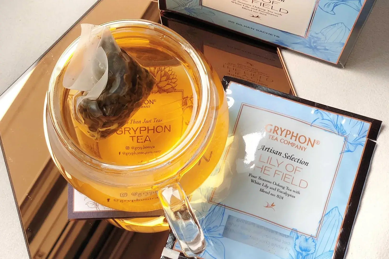 A top-down shot of a clear glass mug filled with amber-colored tea, sitting on a reflective copper-tinted mirror surface that captures its golden glow. Inside the mug, a mesh pyramidal tea sachet floats at the surface, showing visible steeped tea leaves. Lying next to the glass is a light-blue foil tea packet and its corresponding larger outer box for Gryphon Tea Company's Artisan Selection "Lily of the Field," detailed as a Four Seasons Oolong Tea blend with white lily and eucalyptus.