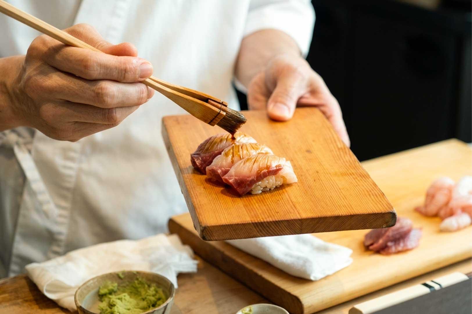 A close-up shot of a sushi chef in Singapore delicately applying a brush of soy sauce glaze over three pieces of fresh nigiri sushi arranged on a traditional wooden serving board, with fresh wasabi and prepared seafood ingredients laid out across the counter.