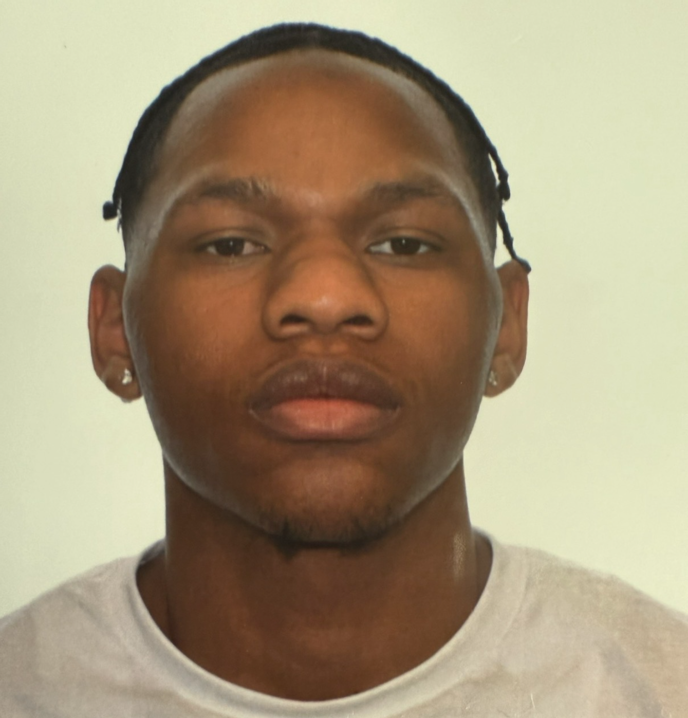 Close-up headshot of a young man with braided hair and small stud earrings, wearing a white crew-neck shirt, photographed against a plain light background — styled as a clean, professional portrait photo.