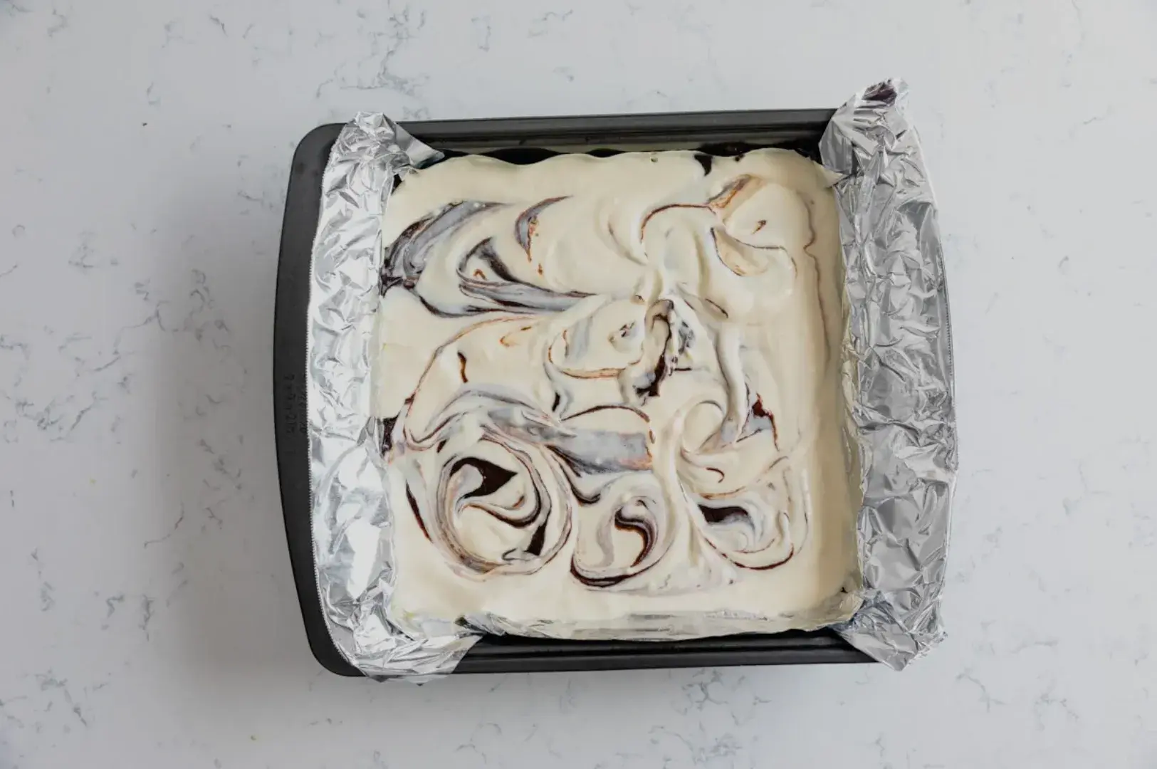 A square baking pan lined with foil, containing a creamy mixture swirled with dark chocolate or fruit puree, sitting on a marble surface.