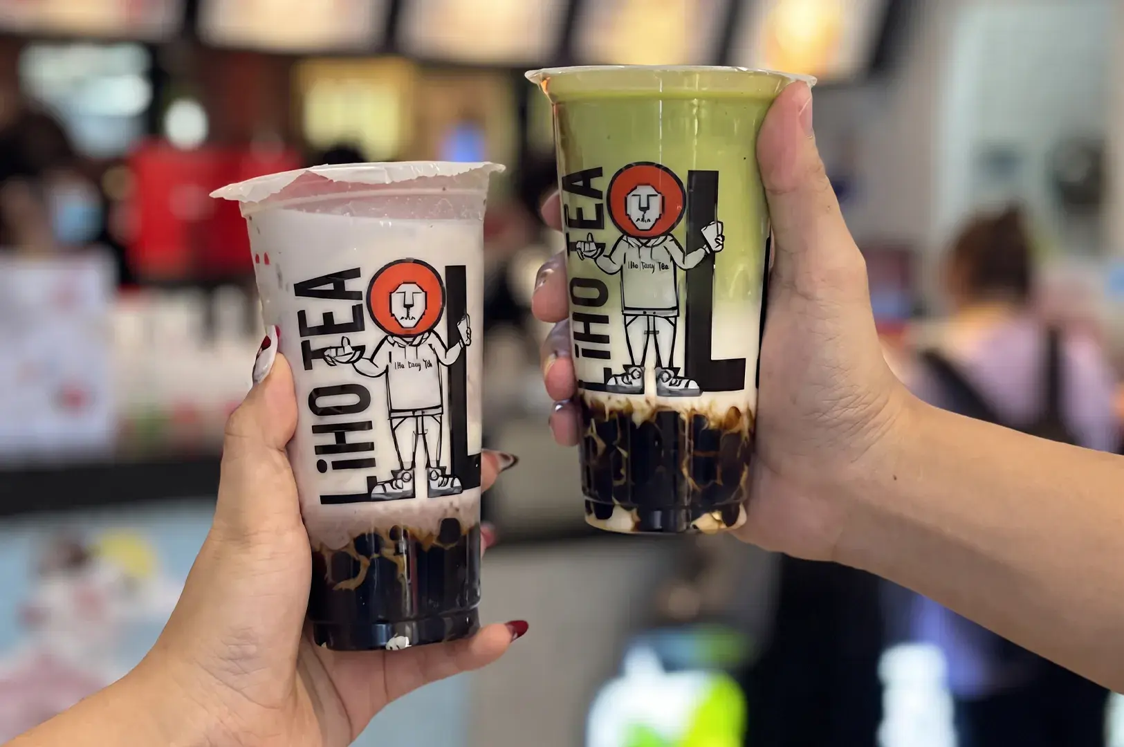 Short Alt Text (Best for general web/social media) Two hands holding up cups of LiHO TEA boba drinks—a taro milk tea on the left and a matcha latte on the right—both with brown sugar tapioca pearls.