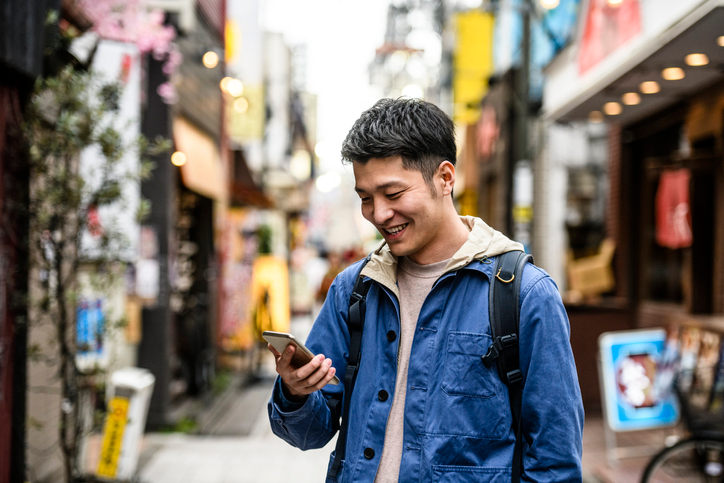 Happy young man in a blue jacket surrounded by shops checking his cell phone. 