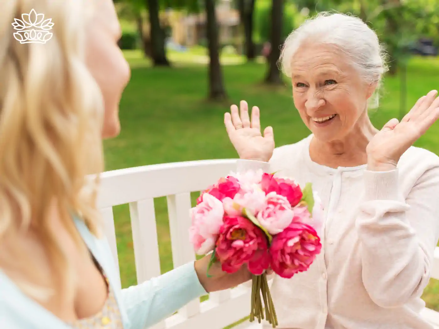 Joyful elderly woman receiving a bouquet of pink peonies from a younger woman on a park bench, expressing happiness and surprise. Fabulous Flowers and Gifts - Thank You Flowers. Delivered with Heart.