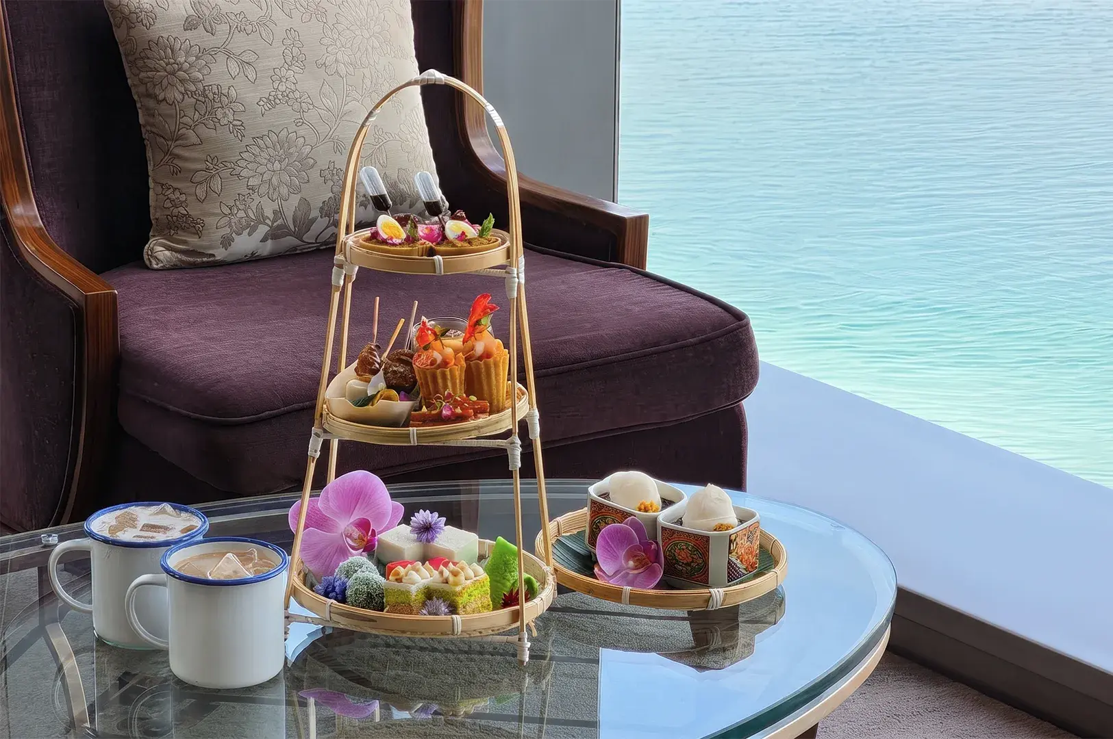 Three-tiered bamboo tray displaying high tea pastries, savory bites, and local desserts on a glass coffee table beside enamel mugs, set against a deep purple armchair and ocean views.