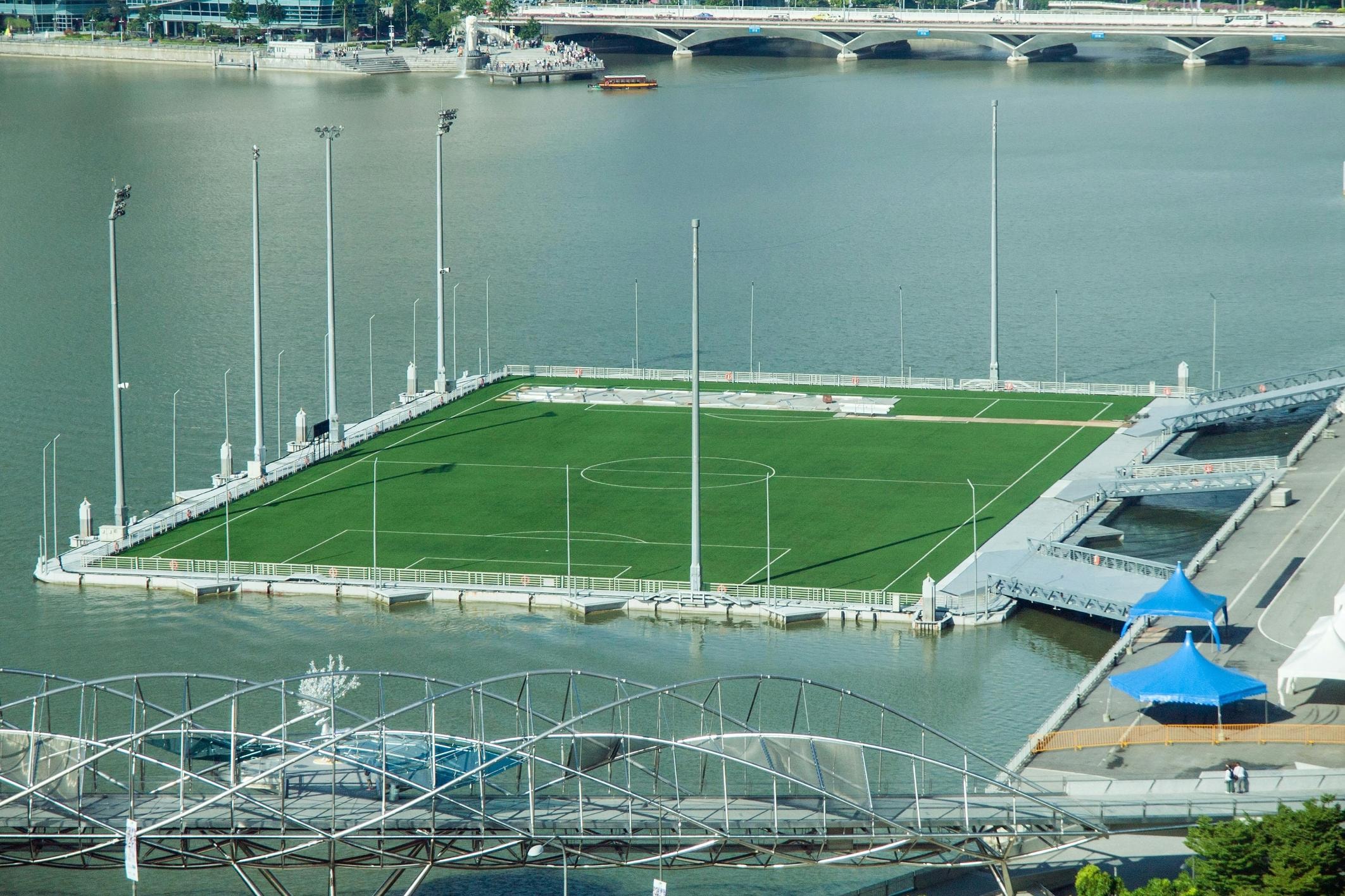 A rectangular green football pitch floats prominently on the water, surrounded by tall floodlight poles and connected to the land by metal walkways. The scene is framed by the intricate lattice of a modern pedestrian bridge in the foreground and a busy highway bridge spanning the river in the background.