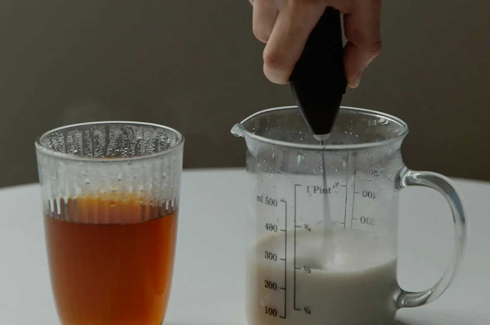 A person's hand is visible from above, holding a black handheld electric frother and lowering it into a clear measuring pitcher filled with a creamy, light-colored liquid. To the left, a separate glass cup filled with dark, amber-colored tea sits on a white surface, suggesting the preparation of a milk tea or latte.