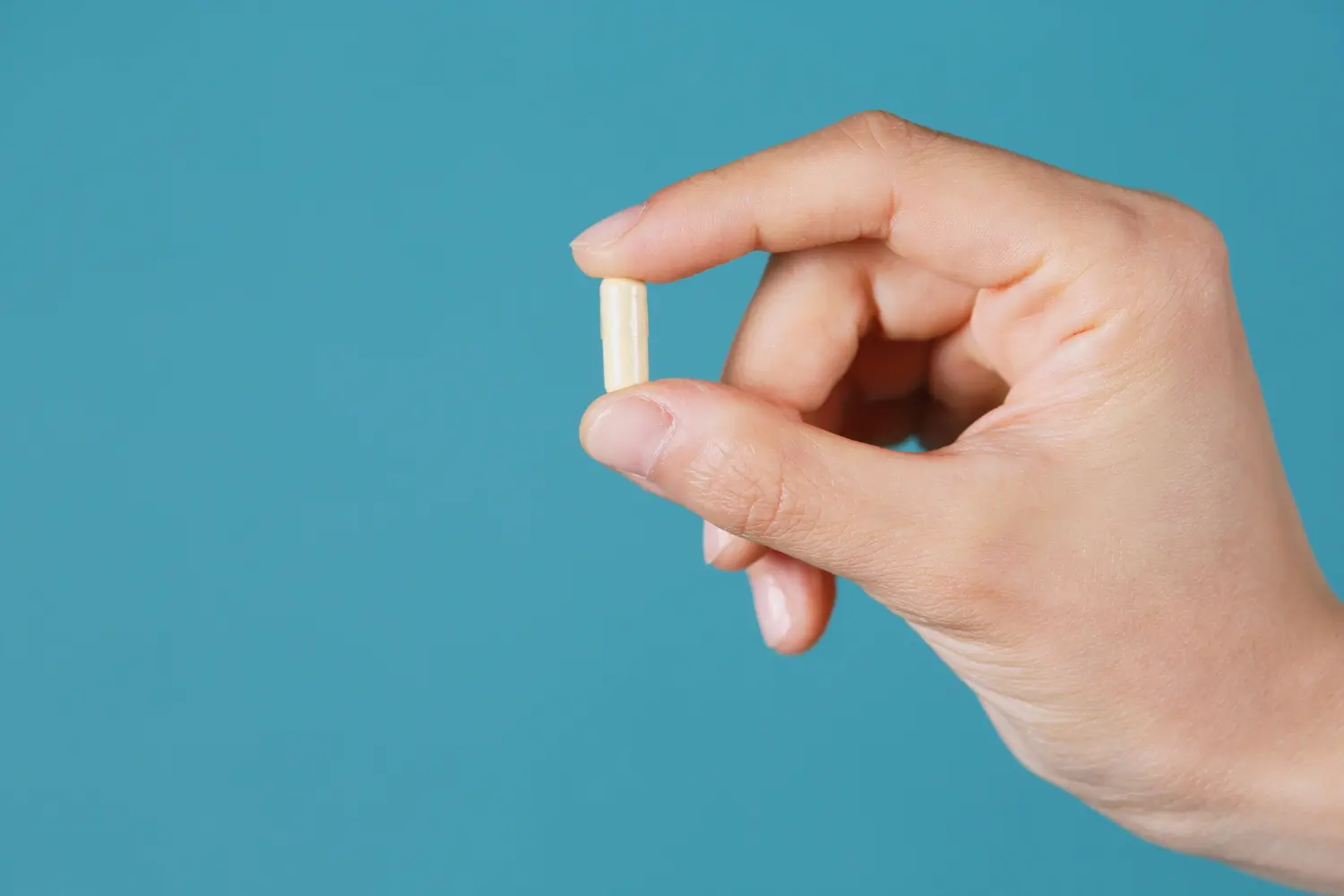 A hand holds a single light-colored lithium orotate capsule between the thumb and index finger against a blue background.