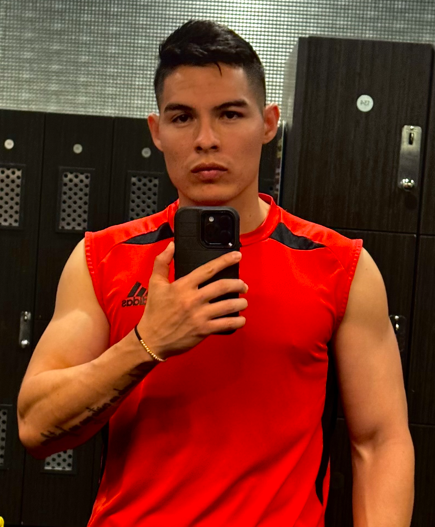 A fit young man takes a mirror selfie in a gym locker room, wearing a red Adidas sleeveless athletic shirt and black shorts, conveying a post-workout or pre-workout confidence — a gold bracelet and a yellow wet floor sign visible in the background.