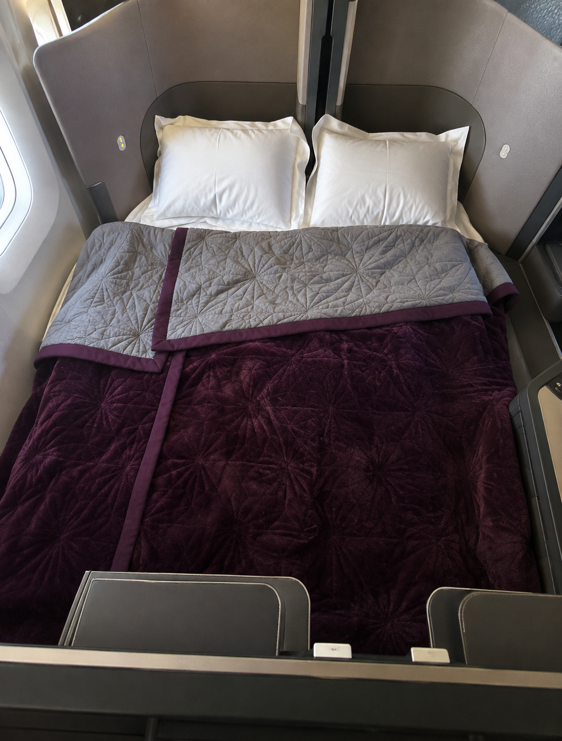 Business class double bed with two white pillows, a deep purple quilt and a grey patterned blanket inside a private aircraft suite.