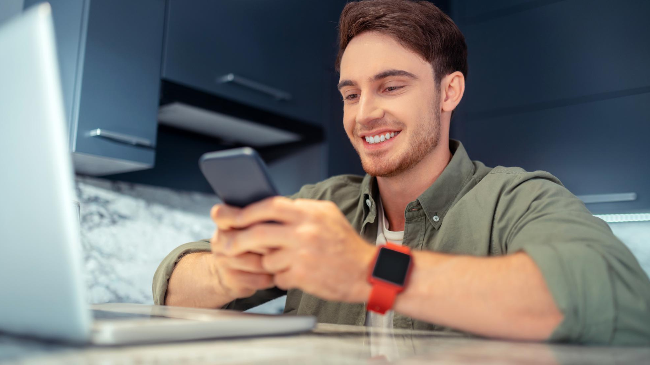 Smiling man in a kitchen using a smartphone with a laptop nearby, illustrating how users can easily receive calls through Ten4’s solution.