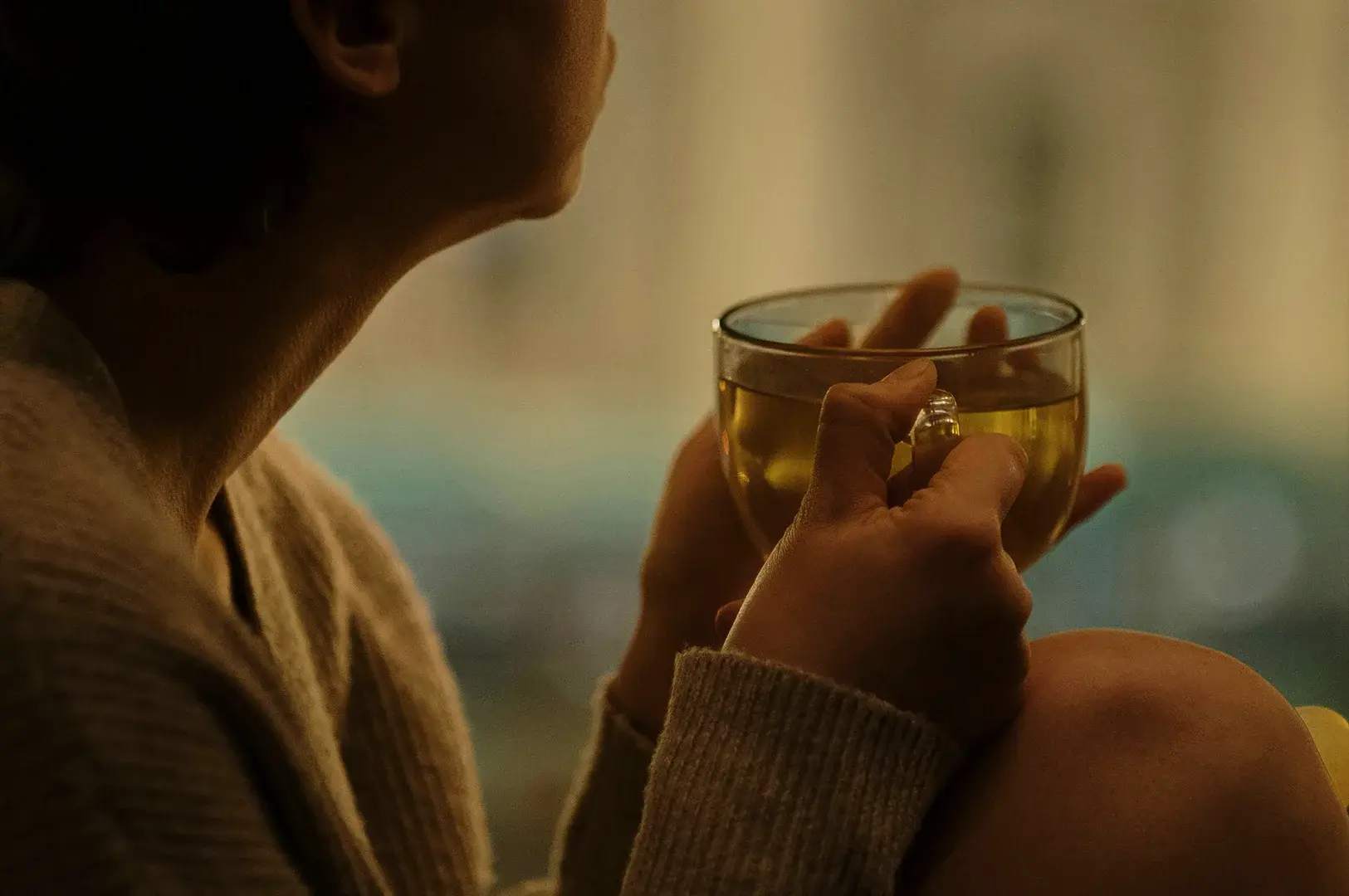 A cozy, close-up shot from behind the shoulder focuses on a person holding a large, clear glass mug filled with a translucent, golden-yellow tea with both hands. The individual wears a thick, textured beige or grey knit sweater, and their hands are wrapped firmly around the warm glass, while the background is softly blurred with warm, ambient lighting, suggesting a relaxed indoor evening setting.