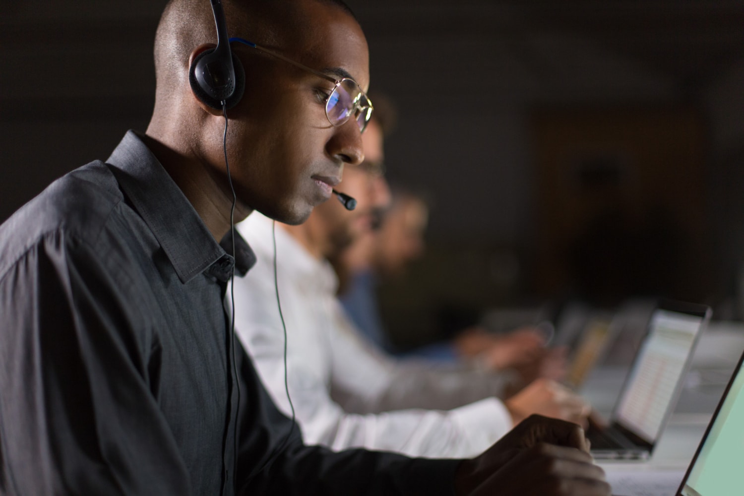 A focused IT professional wearing a headset works alongside colleagues in a dimly lit environment. The image showcases cloud-based telecom, customer communication tools, and managed telecom services.