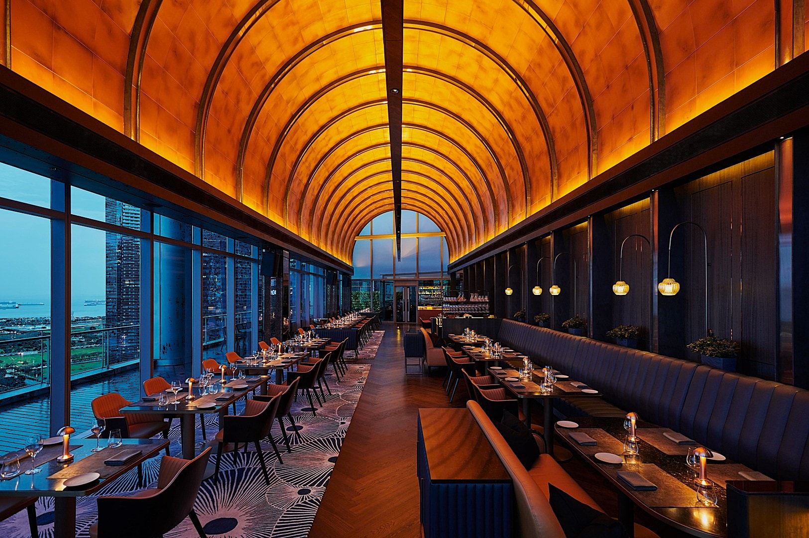 Luxurious restaurant interior with a golden arched ceiling, ambient lighting, and large windows offering a city view. Elegant dining tables and blue seating create a sophisticated atmosphere.