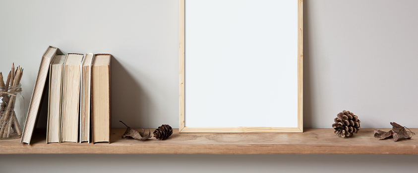 A natural wood shelf. It is holding a blank canvas, some beige books, a glass jar of pencils, and assorted brown leaves and pinecones.