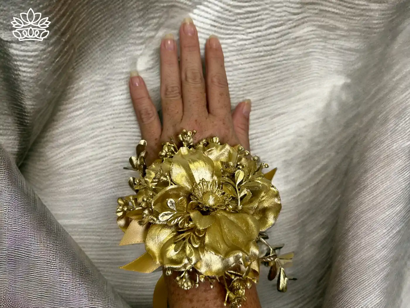A hand elegantly displays a golden floral corsage, adorned with metallic gold flowers and leaves, accentuated by a shimmering ribbon, against a textured silver fabric backdrop. Fabulous Flowers and Gifts - Matric Dance. Delivered with Heart.