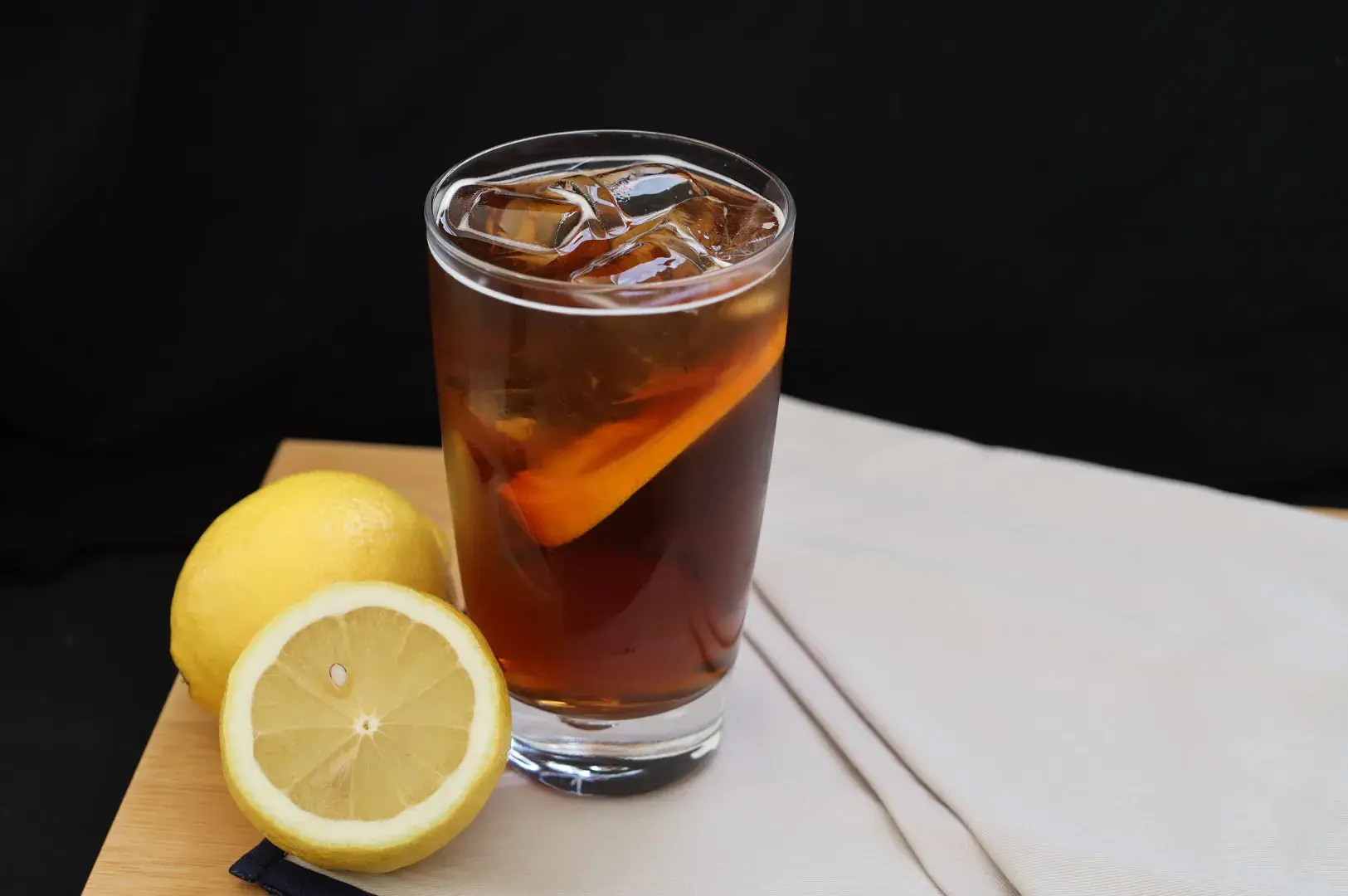 Tall glass of iced lemon tea filled with ice cubes and a lemon slice, accompanied by fresh whole and halved lemons on a wooden surface.