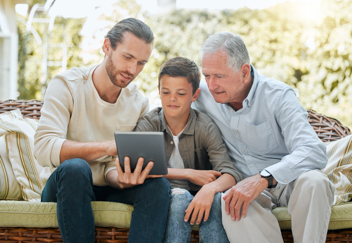 Dad, son, and grandpa sitting outside on a wicker sofa looking at a tablet.
