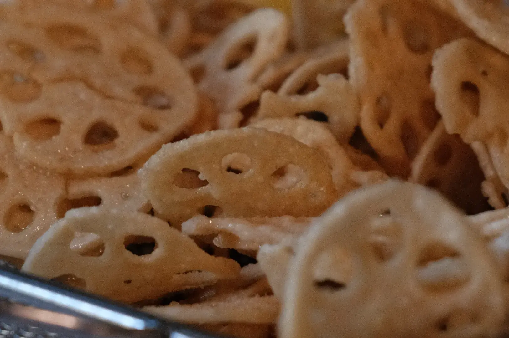 Close-up of golden-brown lotus root chips with distinctive round holes, piled together beside a metal serving utensil; warm, appetizing snack presentation.