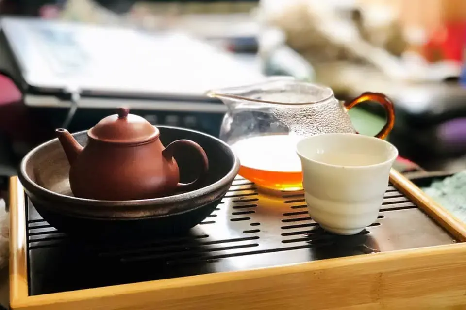 A traditional clay teapot sits nestled in a dark ceramic bowl atop a slotted wooden tea tray. Beside it, a glass pitcher filled with amber liquid and a simple white ceramic cup complete the set.