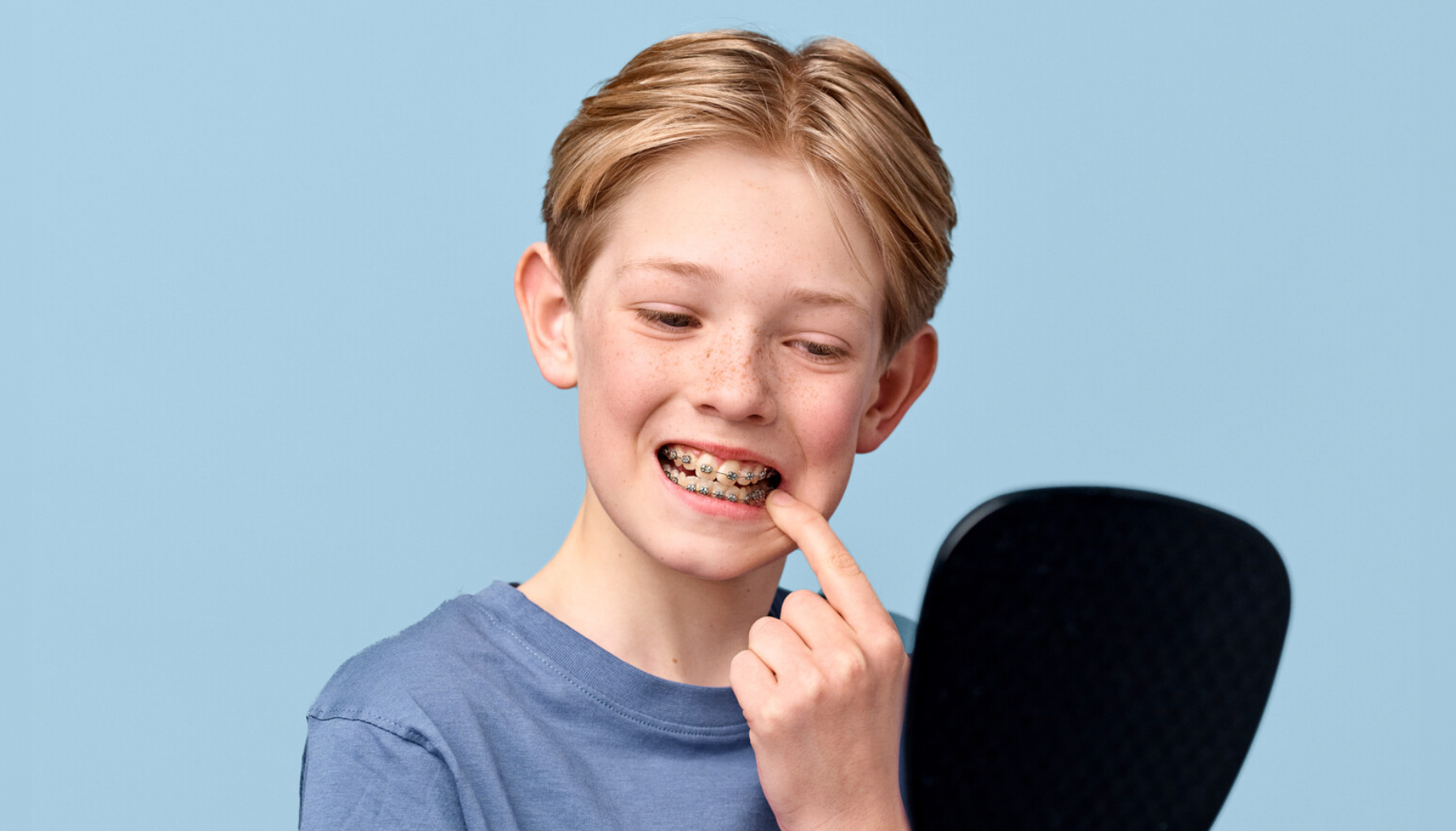 Gutt med tannregulering smiler foran et speil og peker på tennene sine mens han undersøker resultatet.