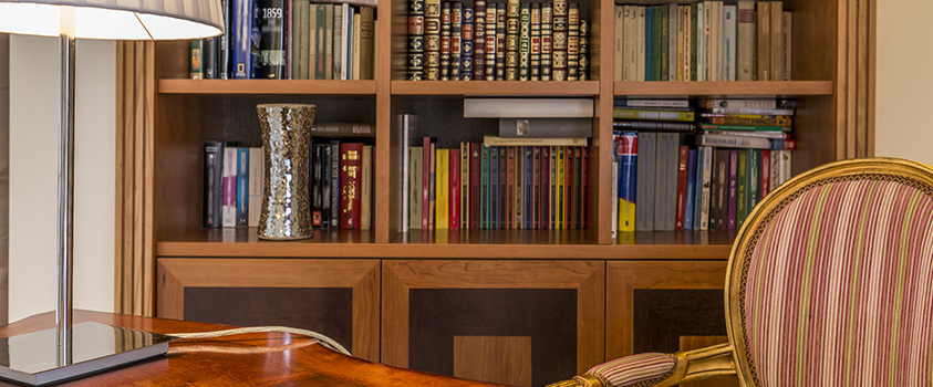 An office with a natural wood grain barrister bookcase, filled with antique books and a silver vase.
