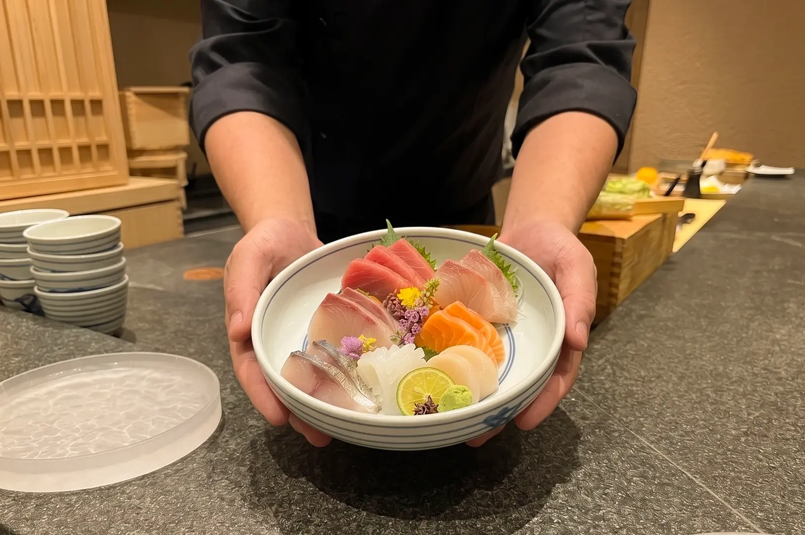 Eye-level close-up of an assorted sashimi bowl being presented across a sushi counter, featuring thick slices of tuna, salmon, white fish, and silver-skinned fish arranged around edible flowers, green leaves, wasabi, and a citrus slice in a white ceramic bowl. Warm restaurant lighting highlights the fresh seafood textures, clean knife cuts, and refined Japanese presentation, with stacked plates and a sushi preparation area visible in the background.