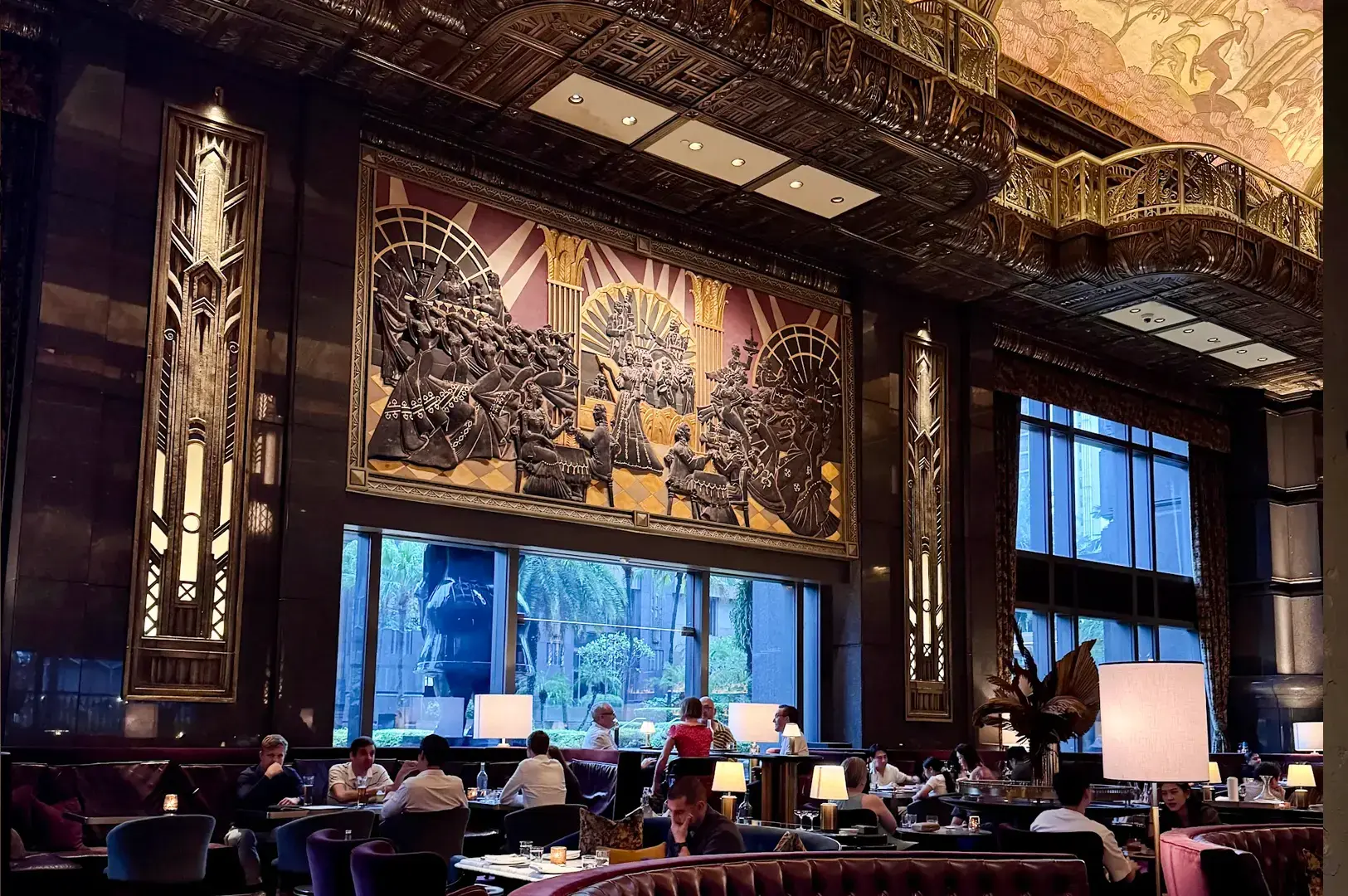 Wide-angle eye-level shot of an upscale cocktail bar lounge with patrons seated at tables, featuring dramatic Art Deco wall panels, sculptural relief artwork, high ceilings, and warm ambient lighting.