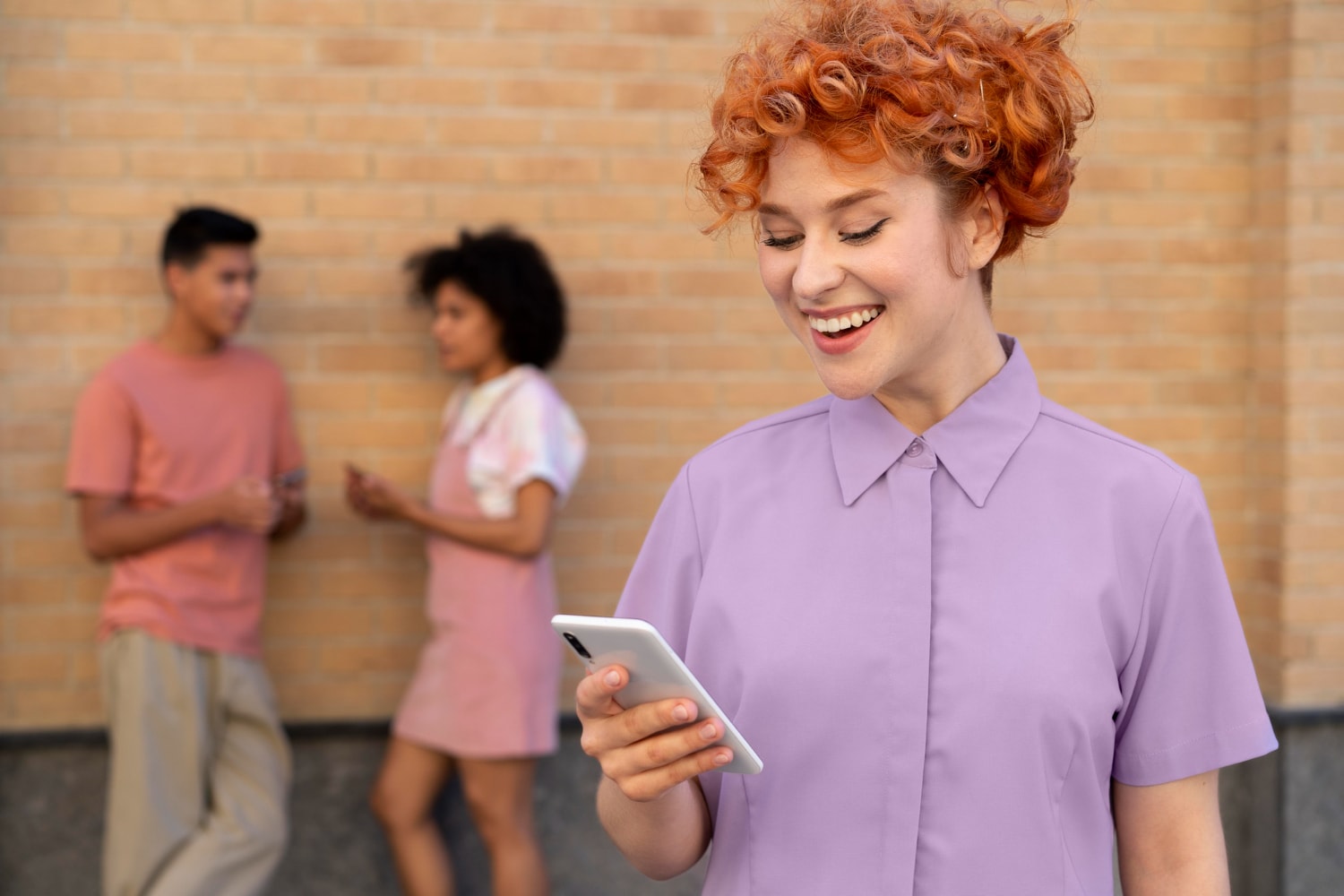 A cheerful woman in a lavender shirt stands in the foreground looking at her smartphone, while two people converse in the background near a brick wall. Her smile reflects the ease of using SMS for business communication and real-time updates.