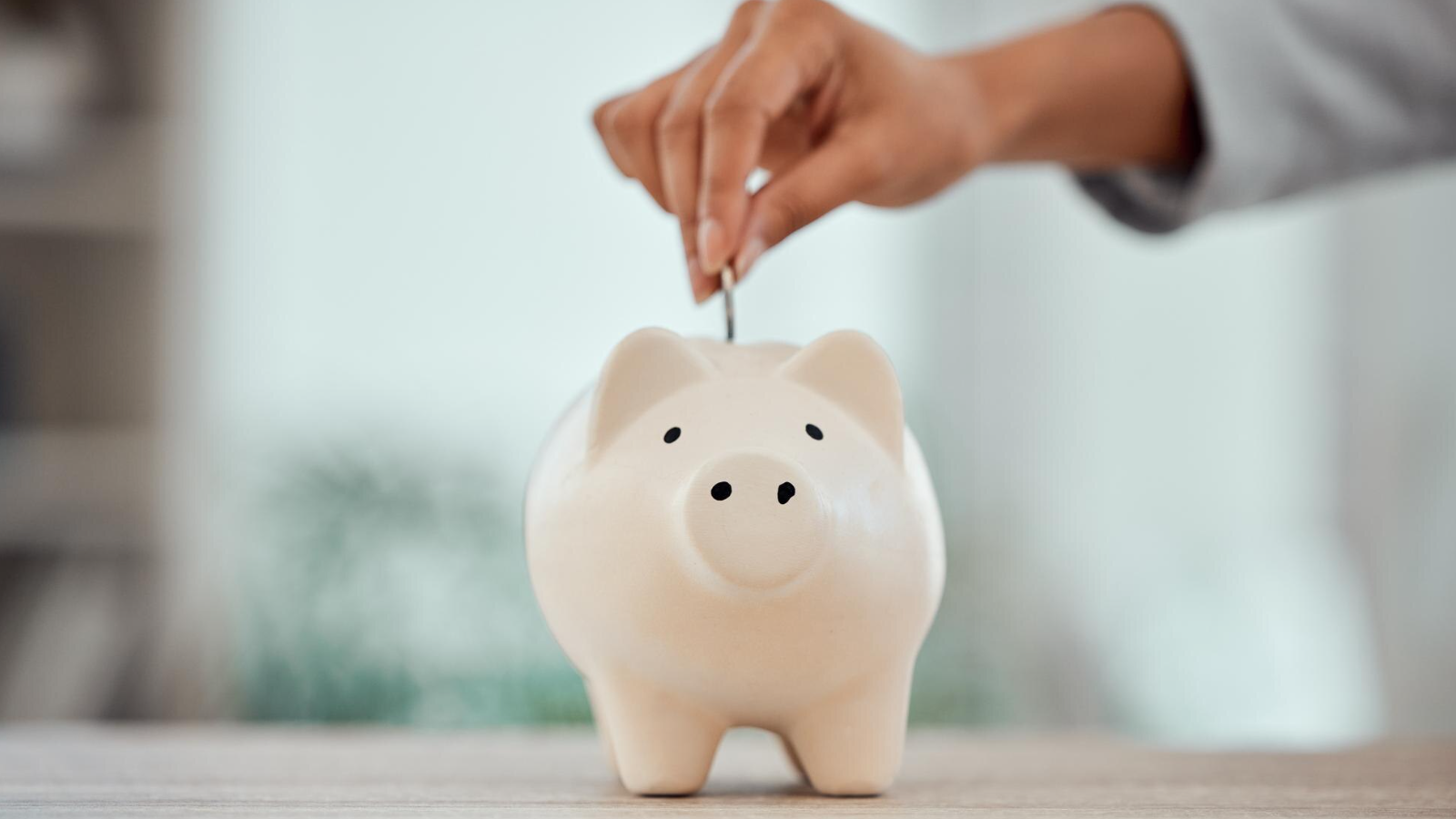 Hand placing a coin into a white piggy bank, symbolizing savings or cost reduction.