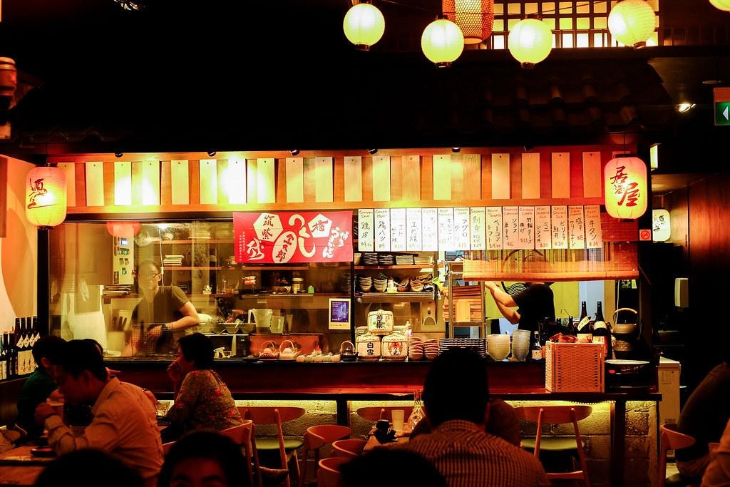  This image captures the bustling, atmospheric interior of a Japanese restaurant, where warm lanterns glow above a busy open kitchen. Customers are seated at tables in the foreground, while staff work behind a service counter decorated with red banners and handwritten menu slats.
