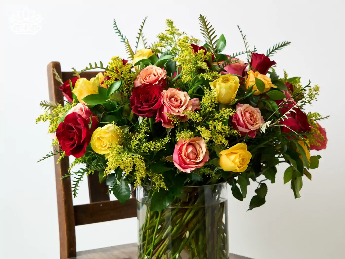Luxurious bouquet of vibrant red, yellow, and pink roses with lush greenery and delicate yellow solidago, displayed in a clear vase on a wooden chair. 