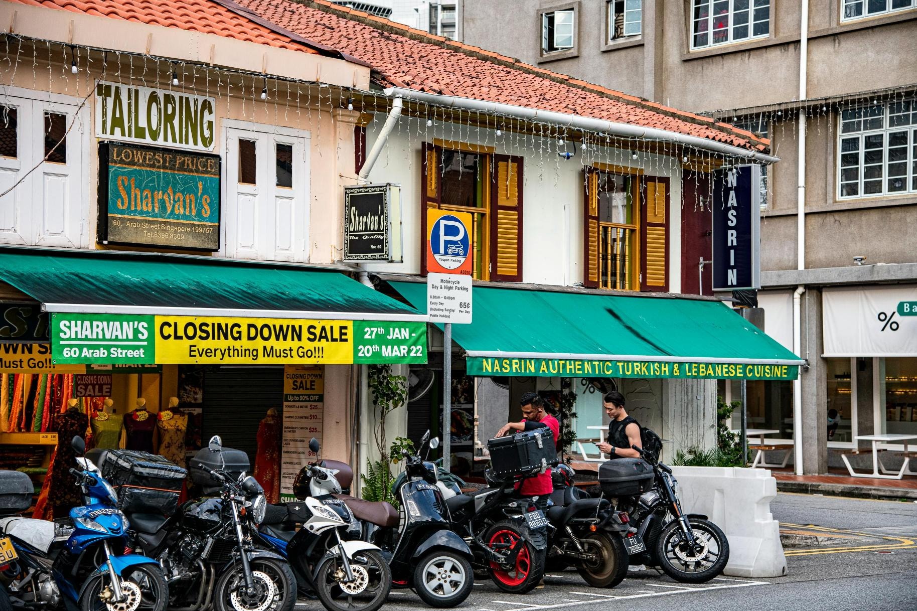 Parked motorcycles line the street in front of traditional shophouses that house a tailoring shop with a prominent "closing down sale" banner and an authentic Turkish and Lebanese restaurant. Two men stand near the bikes, with one adjusting a storage box and the other checking his phone, against a backdrop of tiled roofs and hanging string lights.