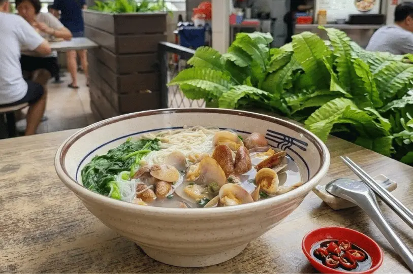 A comforting bowl of clam noodle soup loaded with enoki mushrooms and leafy greens, served with a side of sliced red chilies in soy sauce, alongside lush green potted plants.