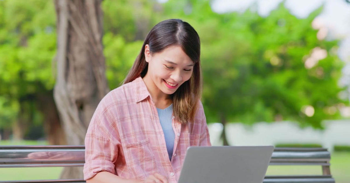 A cheerful woman sitting outdoors on a bench, completing her self employed tax return form on her laptop.