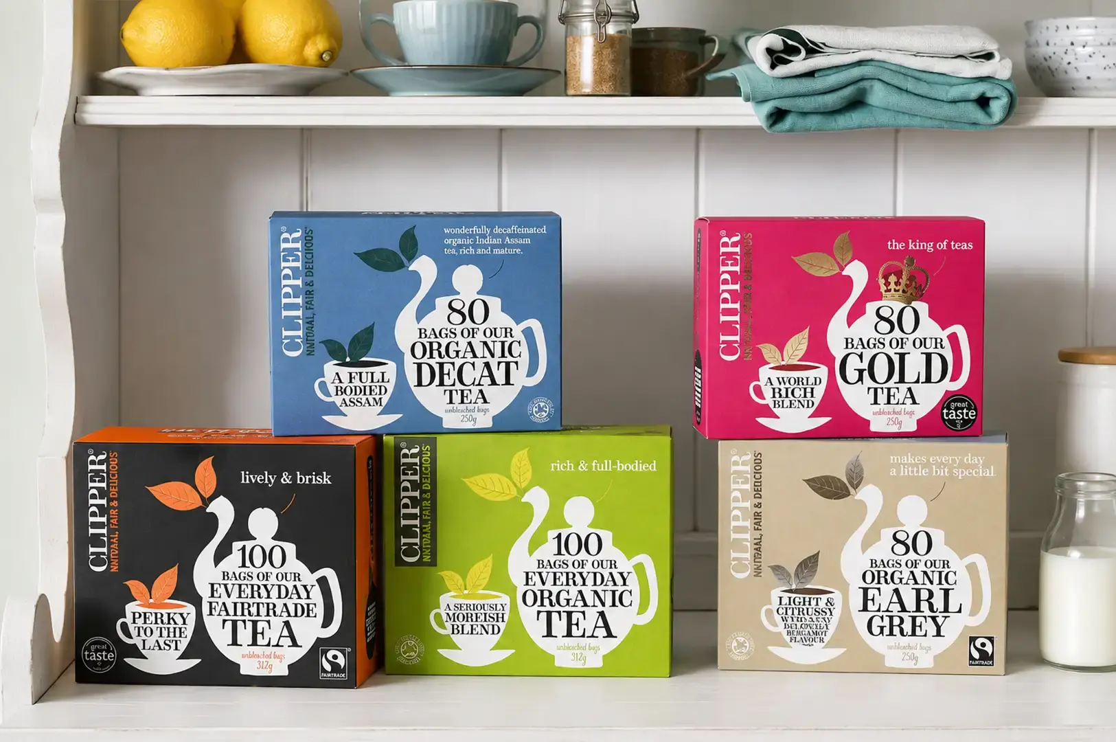 Five colorful boxes of Clipper tea neatly displayed in two rows on a white wooden kitchen shelf, backed by vertical paneling. The top shelf holds blue Decaf Organic Tea and pink Gold Tea boxes, while the bottom shelf showcases black Everyday Fairtrade Tea, green Everyday Organic Tea, and tan Organic Earl Grey Tea boxes next to a glass milk bottle.