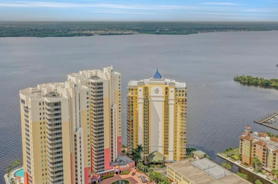 The image depicts a luxury high-rise condominium building located along the waterfront in Fort Myers, surrounded by palm trees. This stunning structure offers immaculate condo units with breathtaking sunset views, providing a truly remarkable living experience for residents.