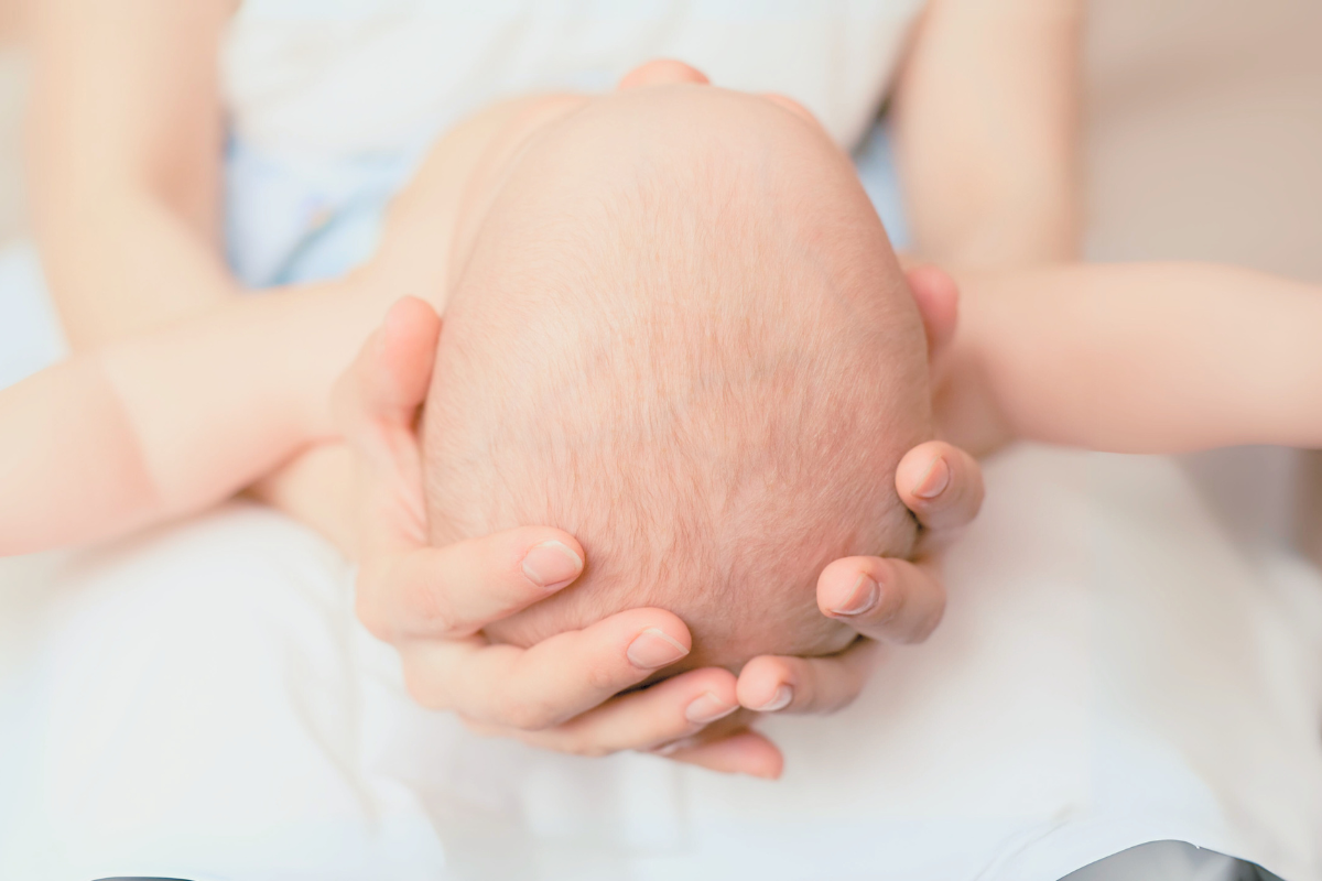 Women holds baby's head in her hands and support the neck. Poppyseed Play