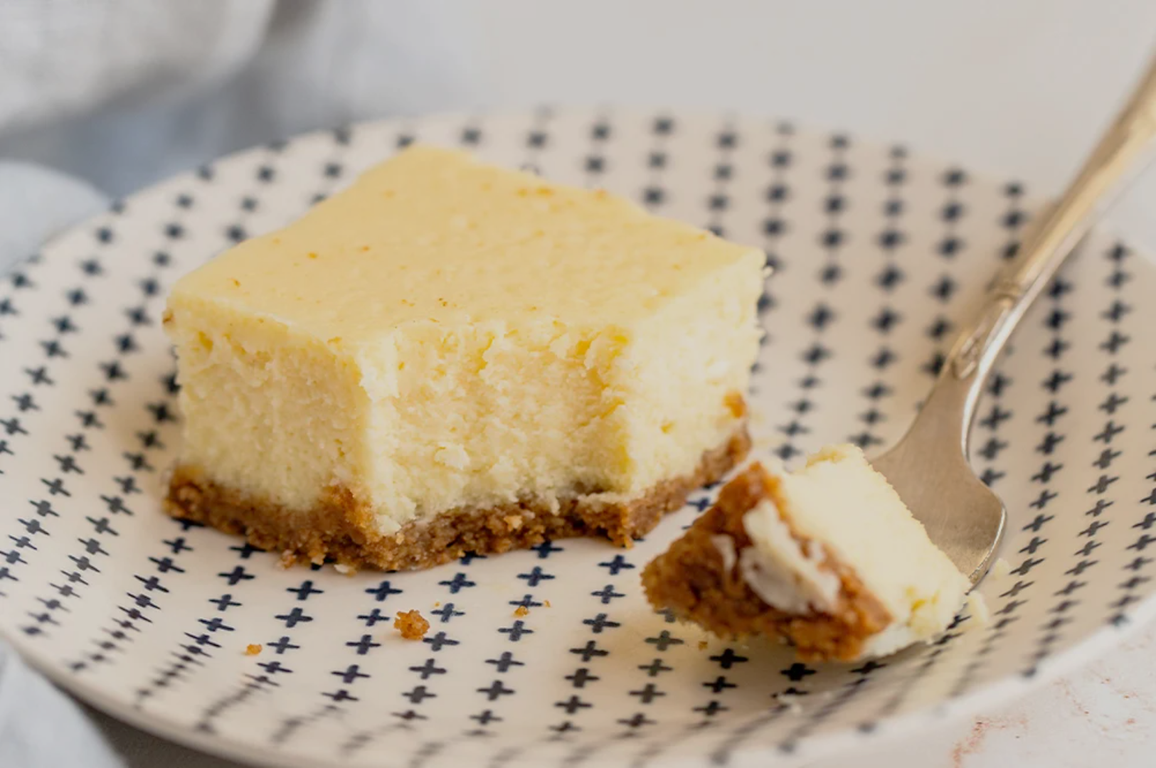 A creamy cheesecake square with a golden-brown crust sits on a patterned plate. A fork with a small piece highlights its texture. Inviting and delicious.