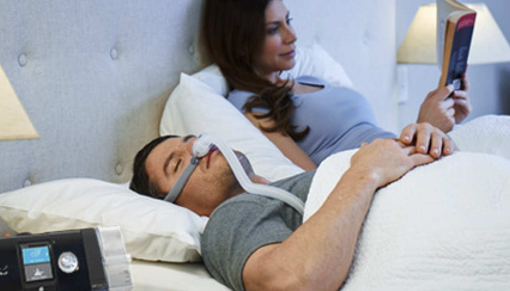 Mann sover med CPAP-maske i sengen mens en kvinne ved siden av leser en bok i et rolig soveromsmiljø.