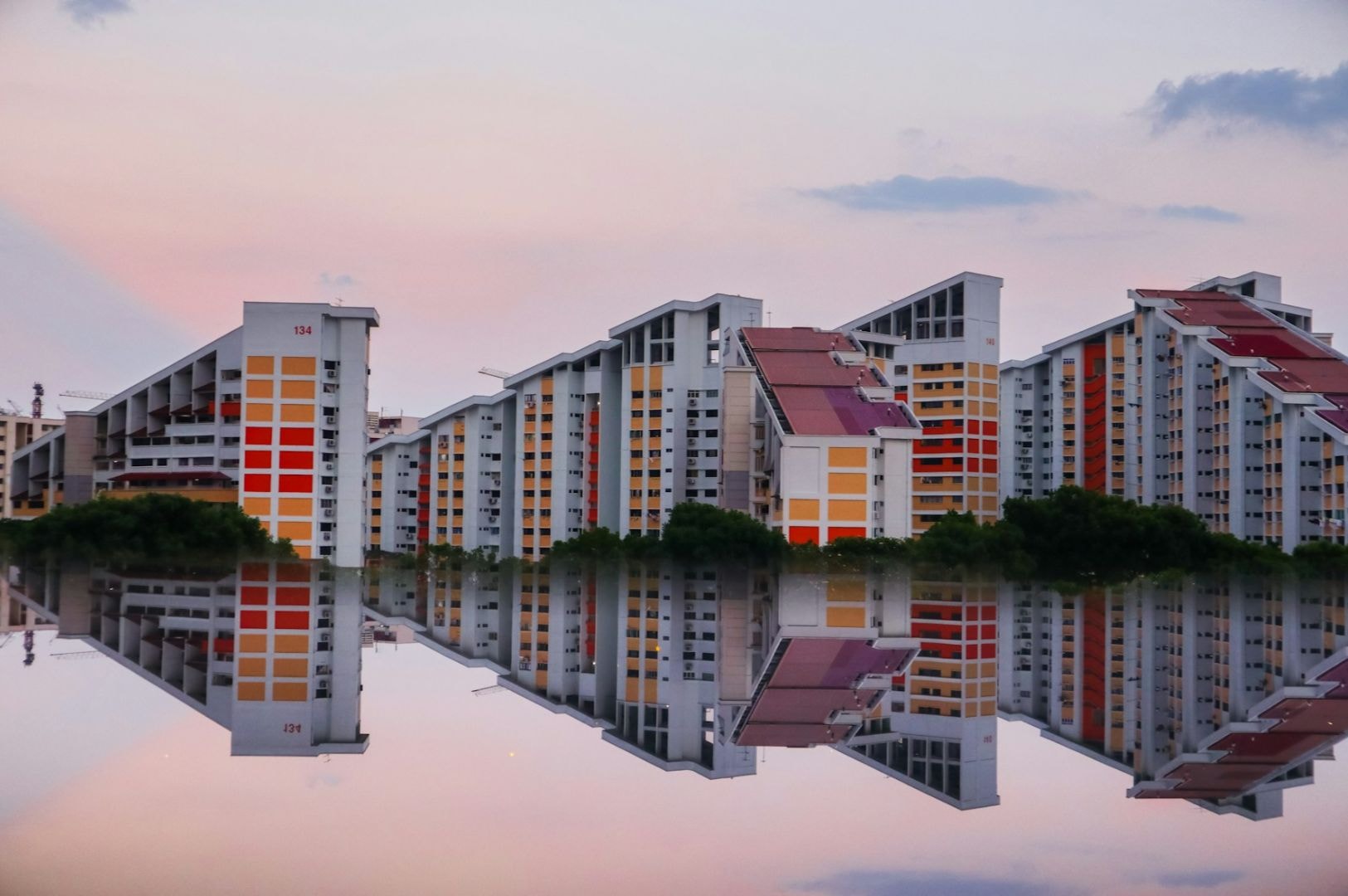 A series of uniquely shaped high-rise buildings with red and orange accents are reflected in a calm body of water, under a pastel pink and blue sky.